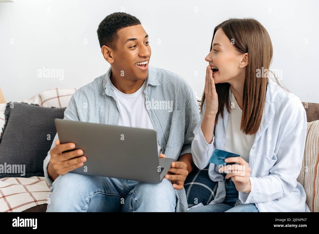 Aufgeregt glücklich stilvolle multirassische Paar verliebt, Ehegatten, sitzen auf dem Sofa in einem Wohnzimmer, mit Laptop und Kreditkarte für Online-Shopping, Lieferung, Blick erstaunt einander, lächelnd Stockfoto