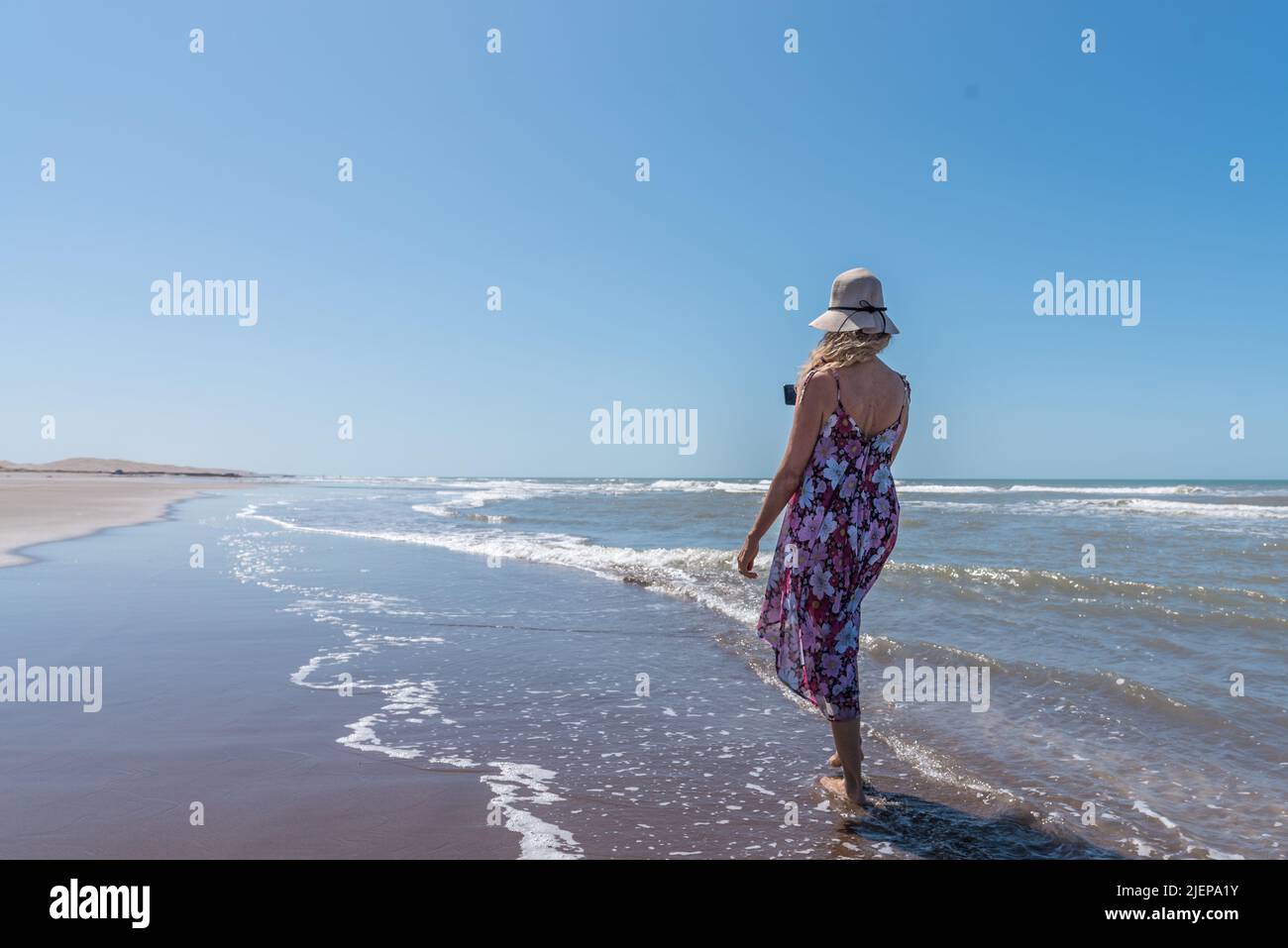 Frau in Sommerkleidung, die barfuß an einem windigen Strand entlang schlendert und mit einem Stockfoto