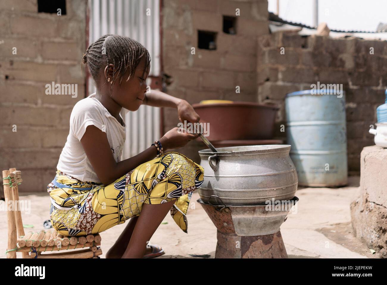 Kleines afrikanisches Mädchen, das vor einem einfachen Kohleofen sitzt, der den Inhalt eines großen Metallköpfen mischt; traditionelle Zubereitung von Speisen Stockfoto