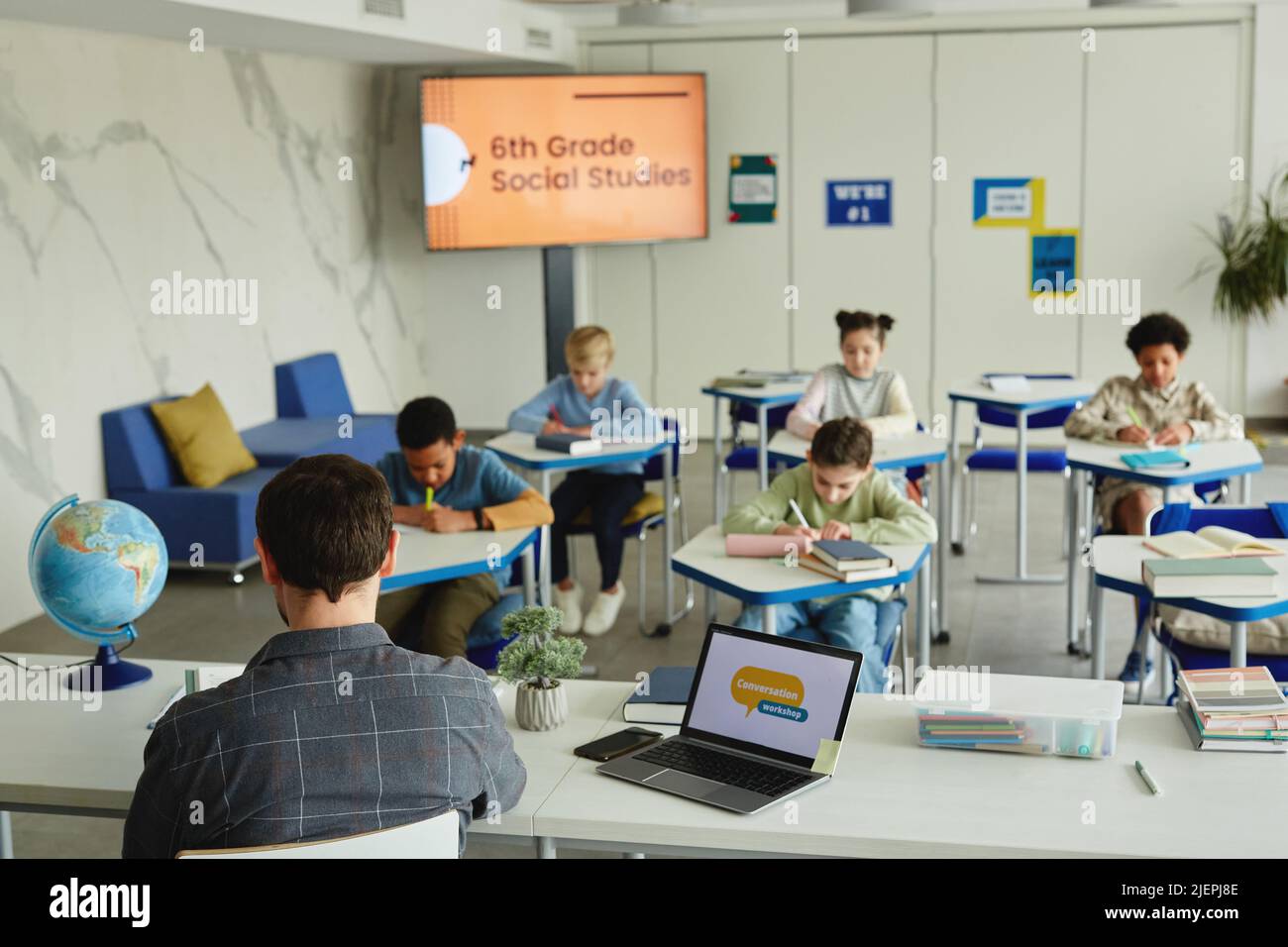 Weitwinkelansicht im Klassenzimmer mit verschiedenen Gruppen von Kindern, die Tests machen, männlicher Lehrer im Vordergrund, Kopierraum Stockfoto