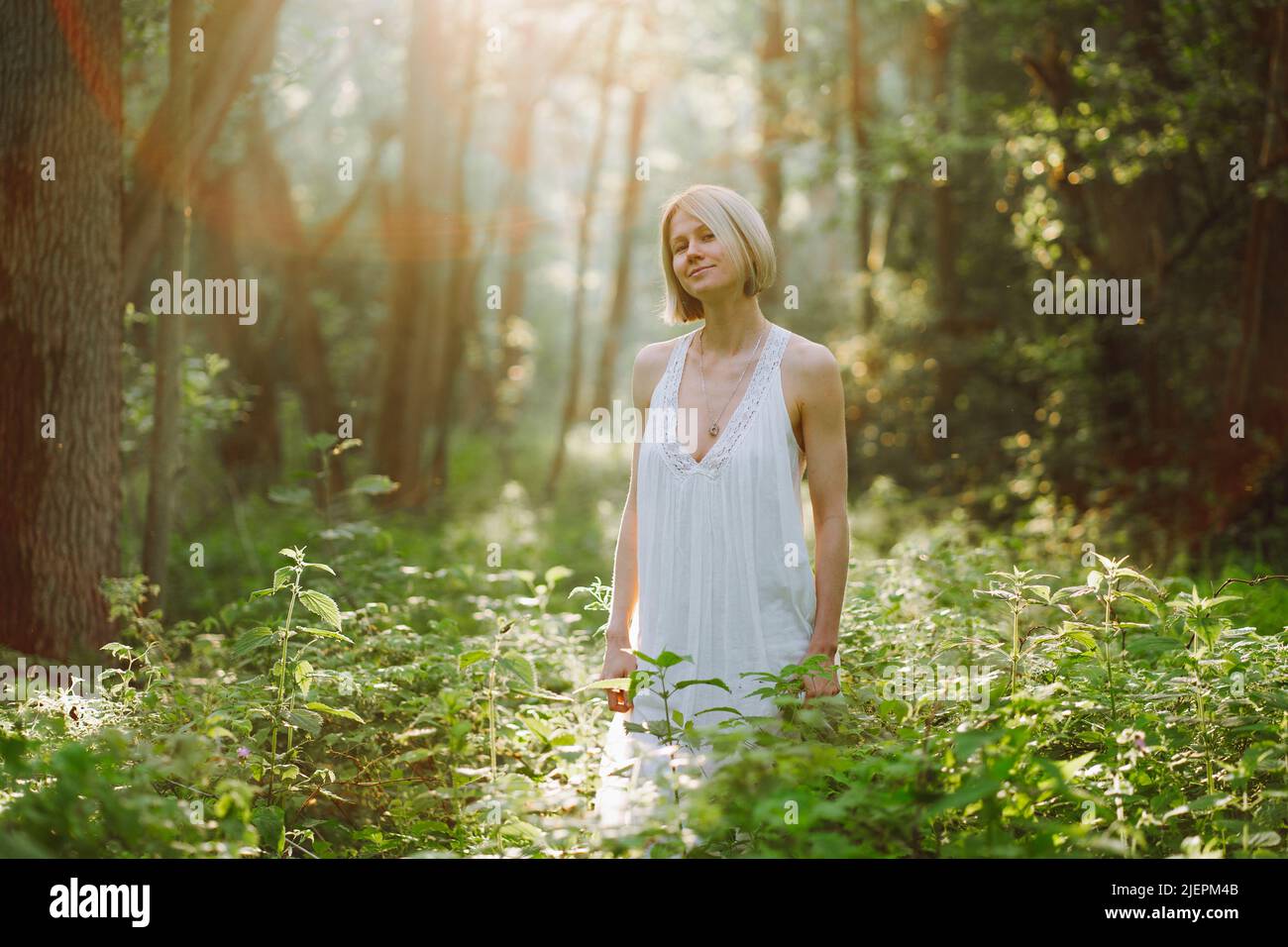Porträt einer friedvollen lächelnden wunderbaren Frau mittleren Alters mit kurzen, hellen Haaren in einem langen weißen Sommersundress, die im Wald zwischen Pflanzen steht Stockfoto