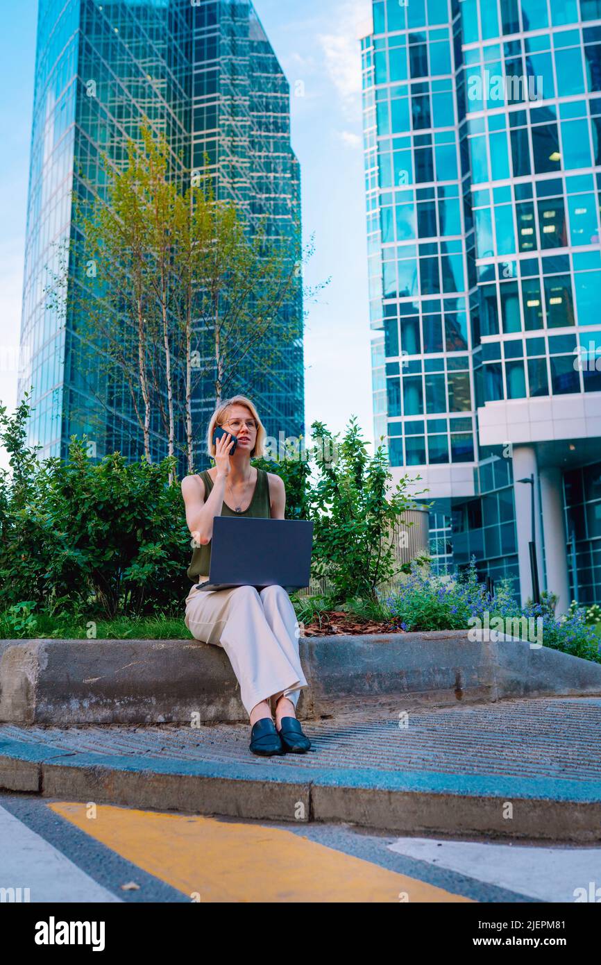 Porträt einer blonden Geschäftsfrau mittleren Alters mit grüner Oberseite, weißer Hose, sitzend auf der Straße im Stadtzentrum in der Nähe von Blumenbetten, Wolkenkratzern, Stockfoto