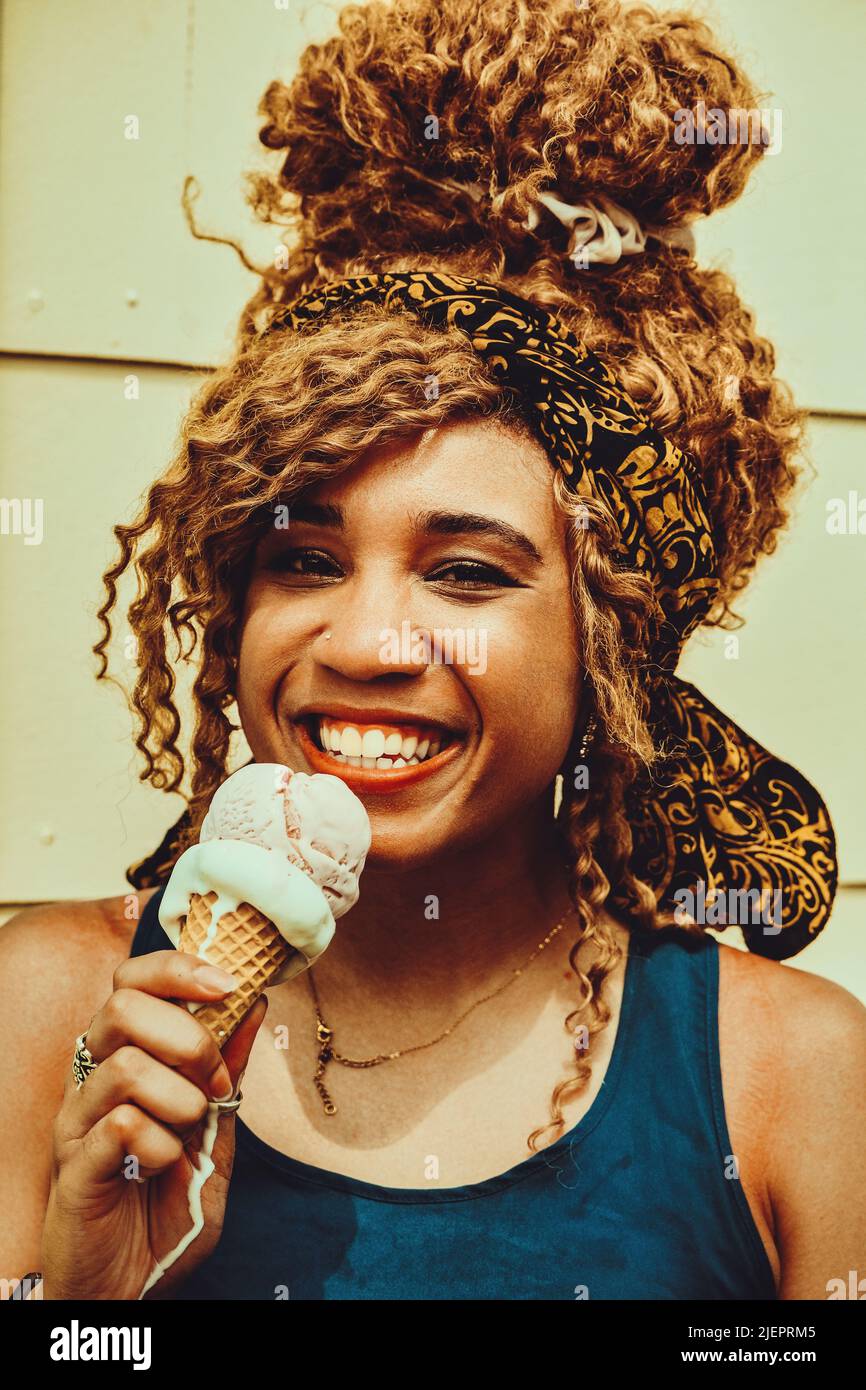 Junge Erwachsene Frau, die im Sommer im Freien Eis isst und mit einem Lächeln auf die Haare lächelt und die Kamera anschaut Stockfoto