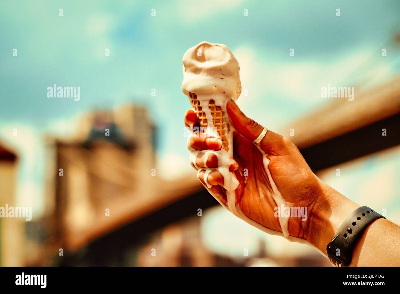 Schmelzender Vanilleeis-Kegel auf der Hand gegen blauen Himmel und Brooklyn Bridge im Hintergrund Stockfoto