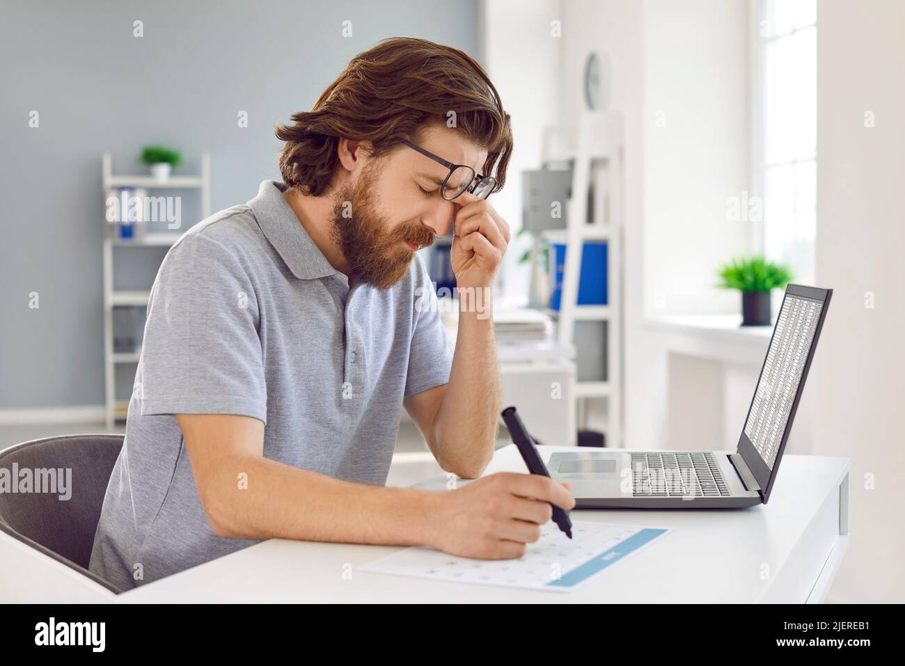 Der müde Mann ist überarbeitet und hat Schmerzen in den Augen, wenn er im Büro am Laptop arbeitet. Stockfoto