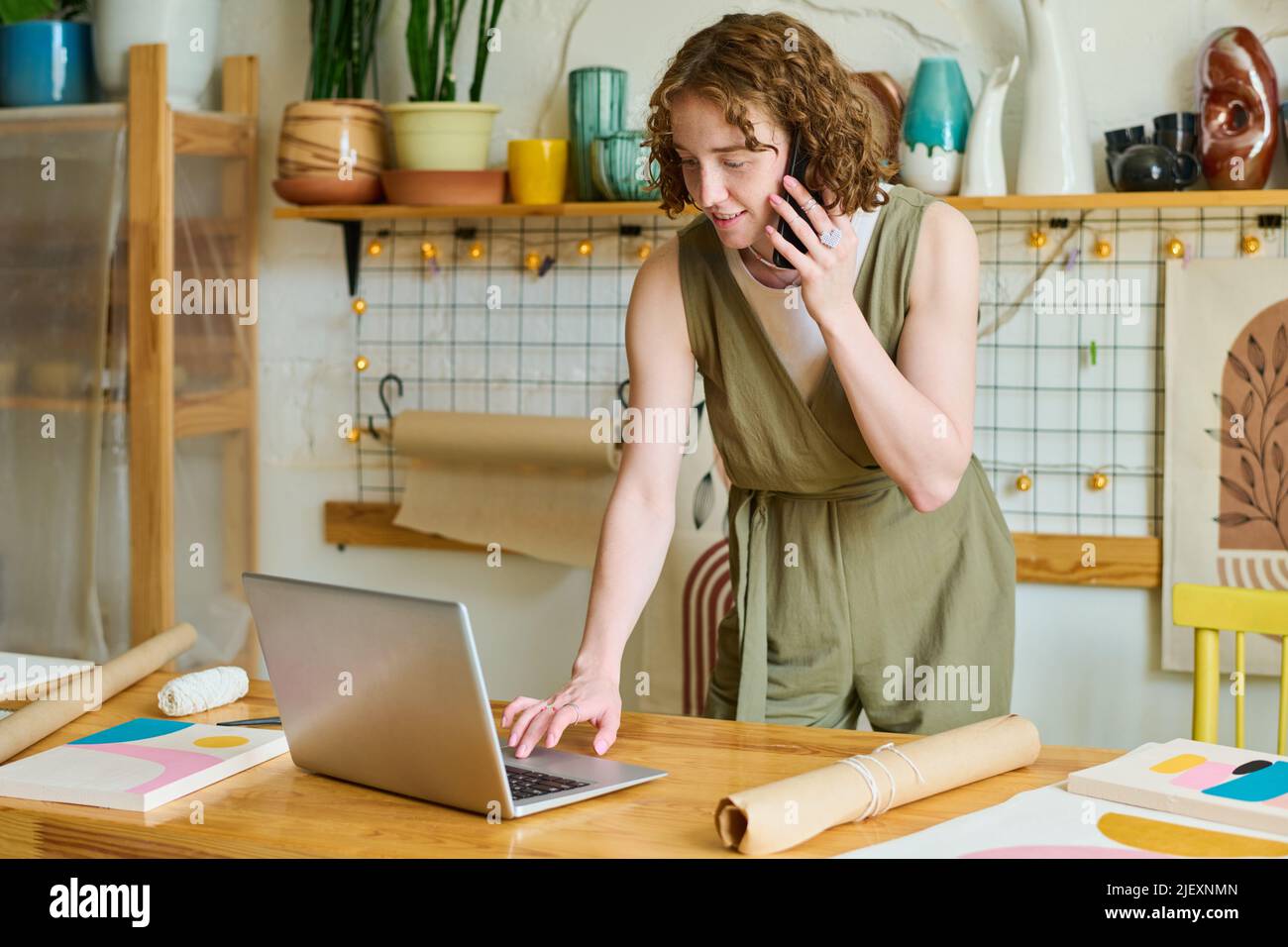 Junge lächelnde weibliche Besitzerin des Kunsthandwerks, die sich mit dem Laptop über den Tisch beugte und Informationen über den Auftrag des Kunden überprüfte Stockfoto