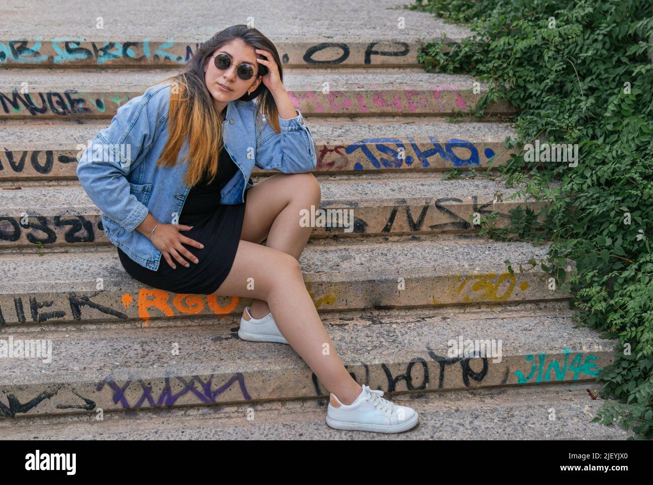 Eine nette lateinerin mit Sonnenbrille und ihrer linken Hand, die ihren Kopf berührt, sitzt auf einer Graffiti-Treppe Stockfoto