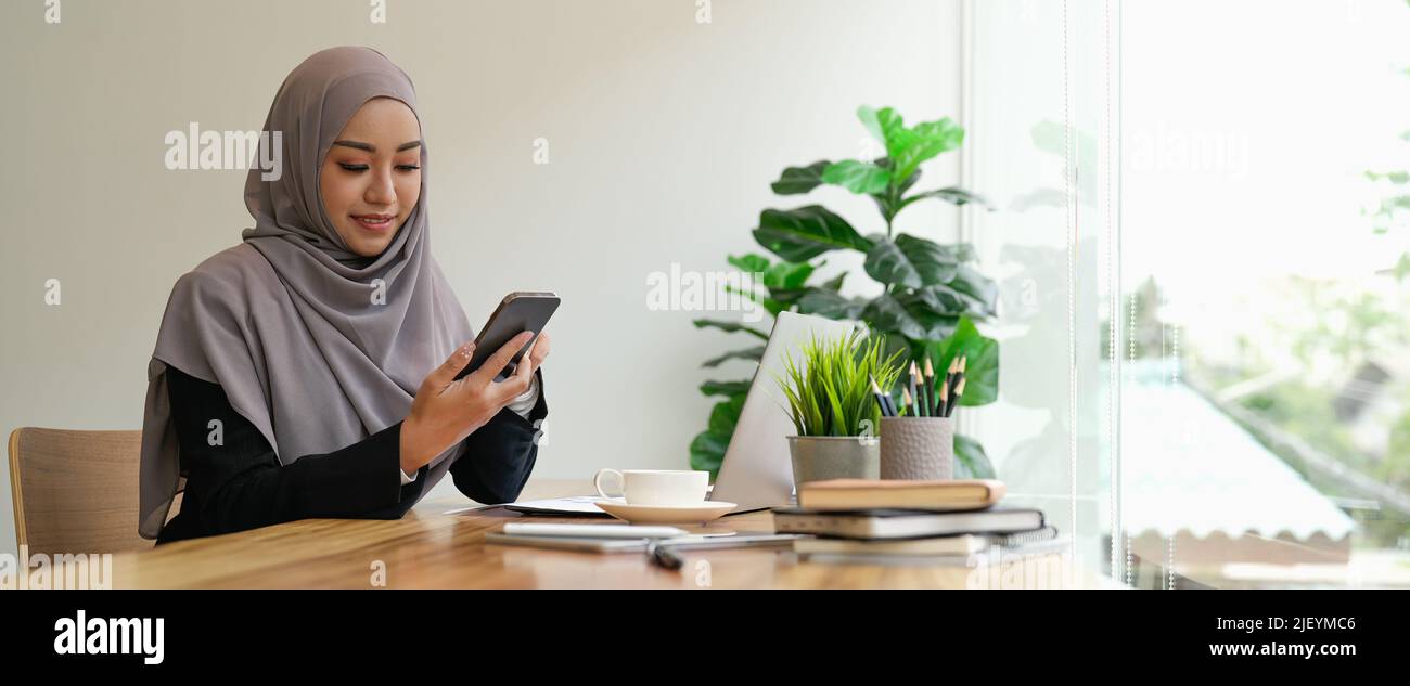 Glückliche asiatische muslimische Frau, die Mobiltelefon in ihrem Büro hält. Stockfoto