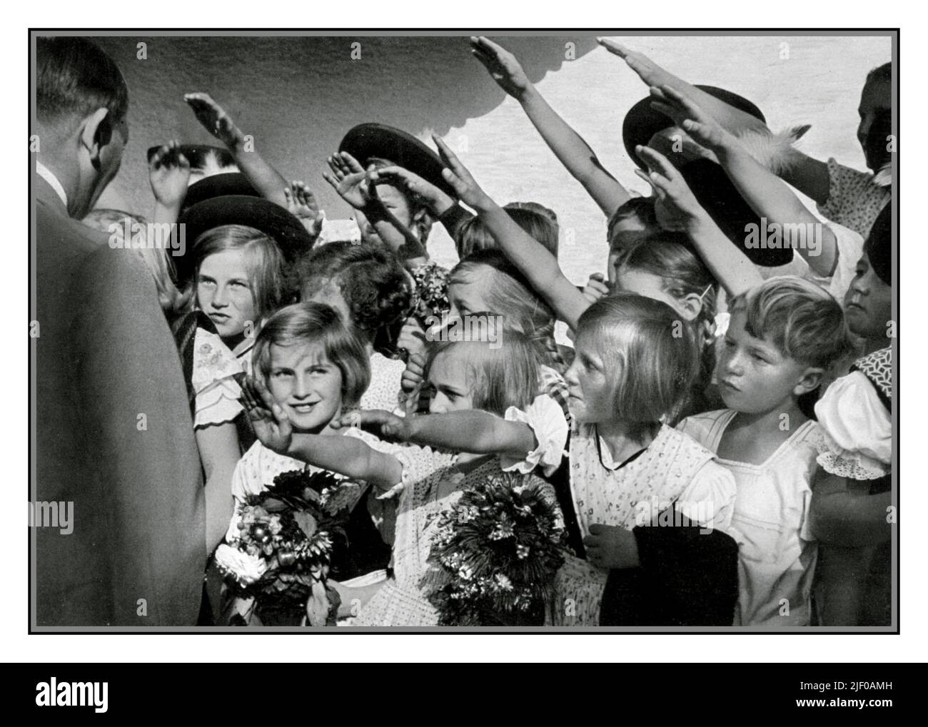 Adolf Hitler 1930er mit einer Gruppe glücklicher junger blonder arier Mädchen und Jungen Kinder mit Blumen, die ihn mit dem Nazi Heil Hitler Salut begrüßen. Nazideutschland 1936 Stockfoto