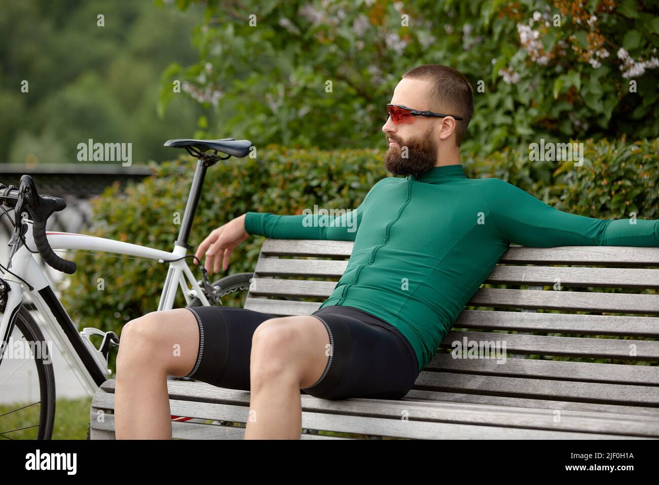 Stilvoller, hübscher Fahrradfahrer ruht auf einer Holzbank in der Nähe des Fahrrads im Sommerpark. Stockfoto