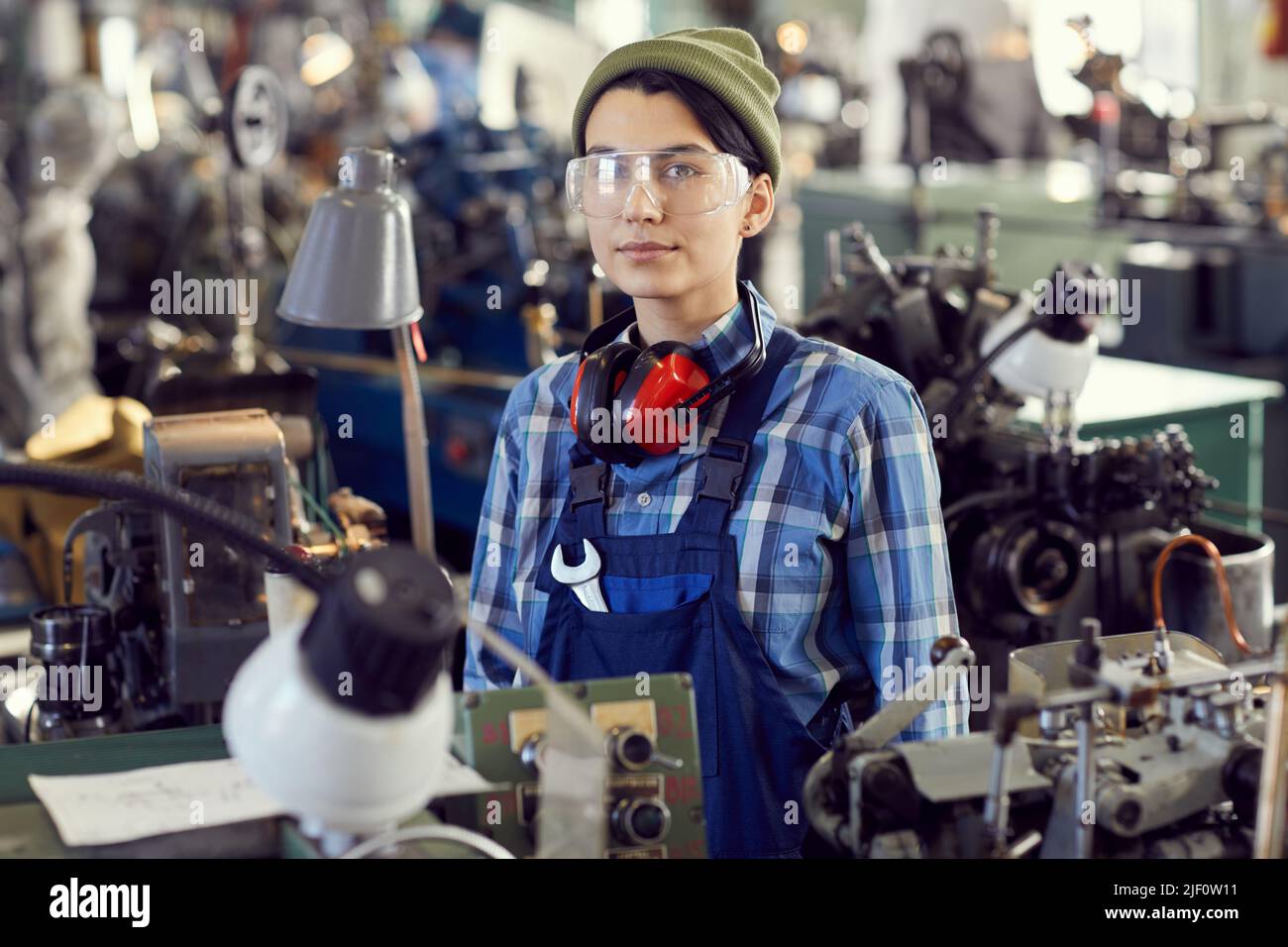 Porträt eines ernsthaften Hipster-Mädchens in Hut und Schutzbrille, das an der manuellen Drehbank steht und diese repariert Stockfoto