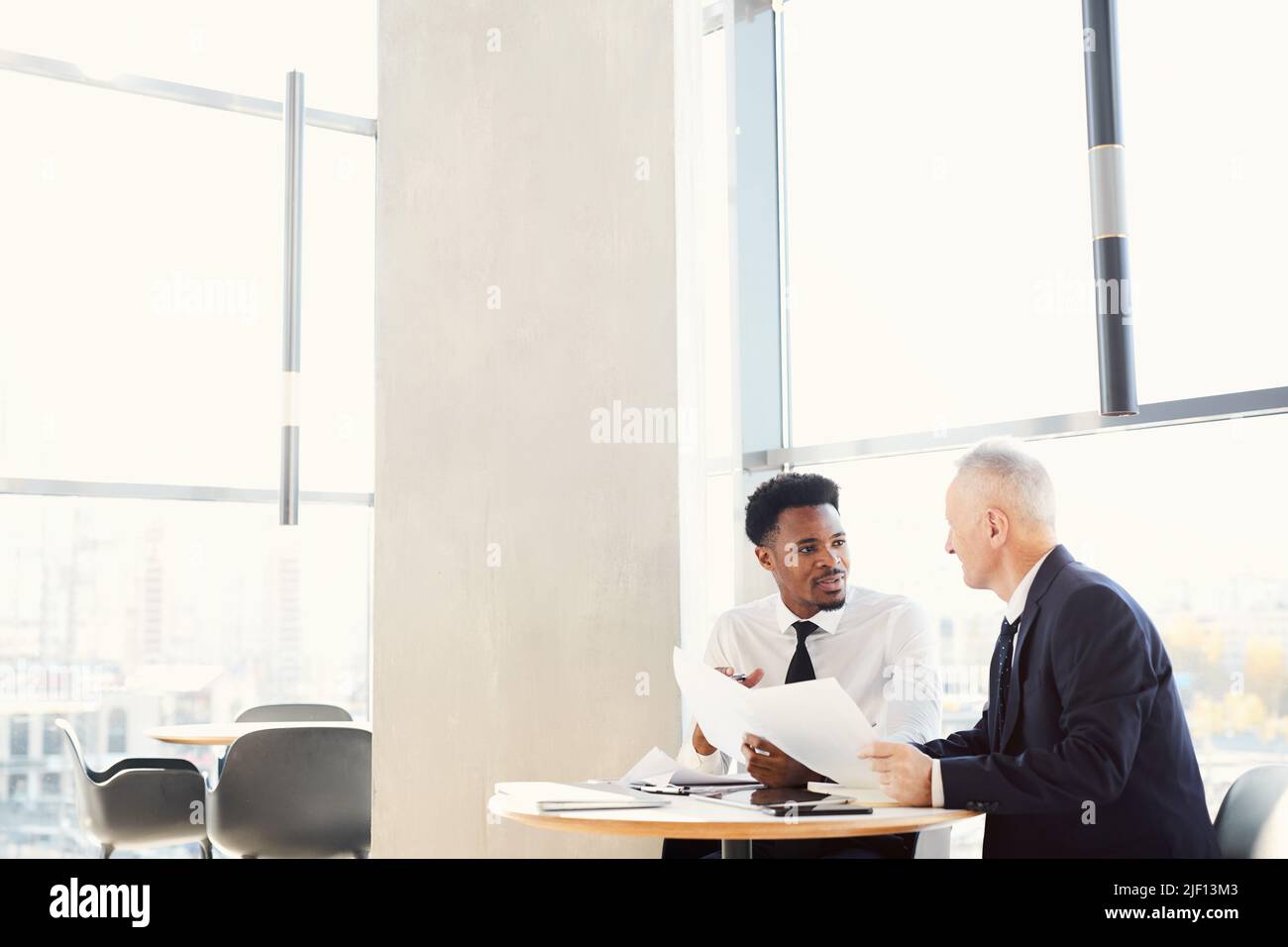 Zwei multiethnische Geschäftsleute in formellen Outfits sitzen im modernen Büro am Tisch und machen gemeinsam Schreibarbeit Stockfoto
