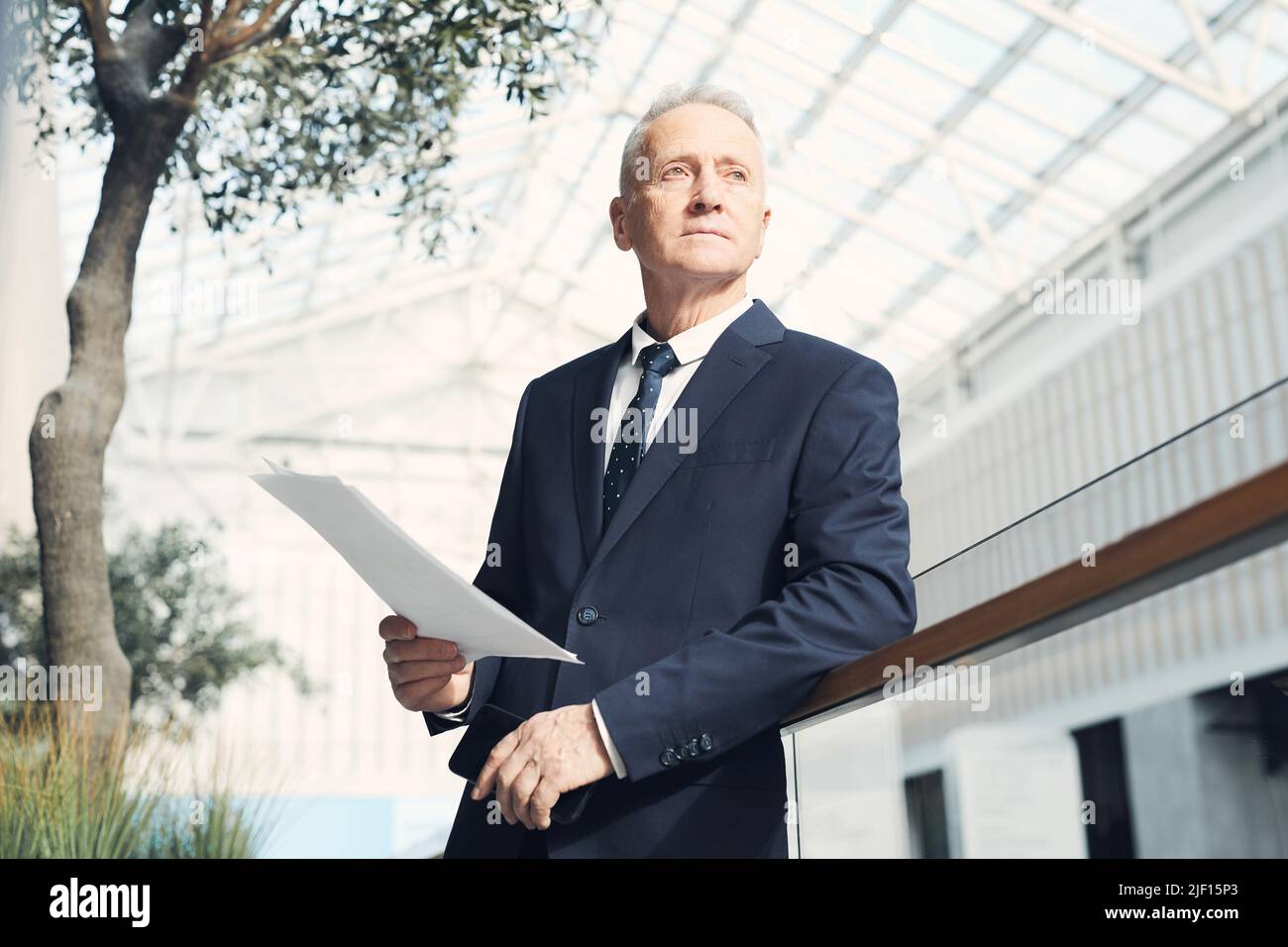 Ernst inspirierter, reifer Finanzier in schwarzem Anzug, der sich in der modernen Lobby auf Geländer stützt und in die Ferne blickt, während er Papiere analysiert Stockfoto