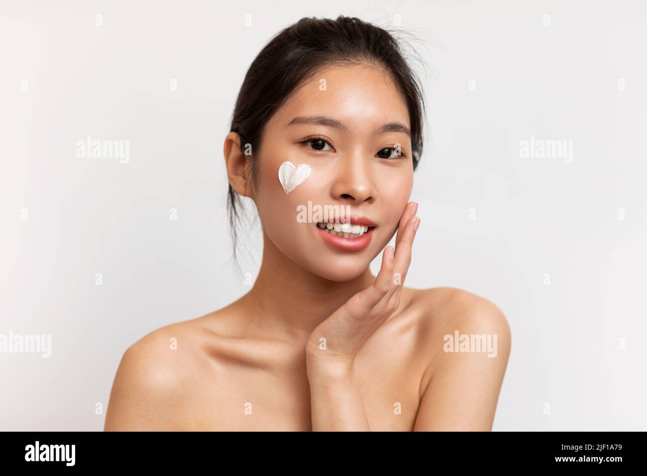 Konzept der Jugendpflege. Junge koreanische Dame mit herzförmiger Feuchtigkeitscreme auf dem Gesicht, die auf weißem Hintergrund posiert Stockfoto