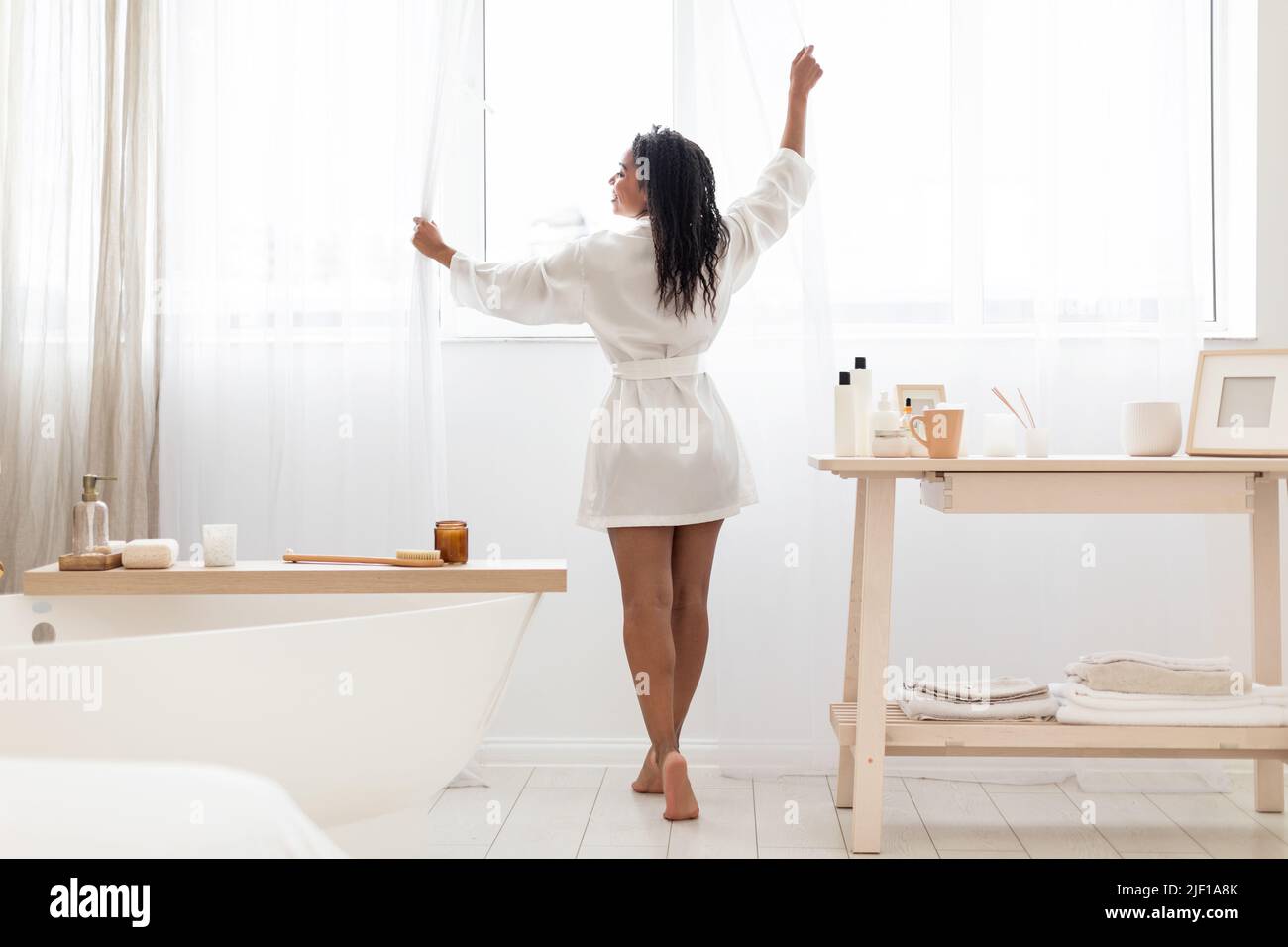 Morgendliche Routine. Glückliche schwarze Frau in Seidengewand, die Vorhänge im Badezimmer öffnen Stockfoto