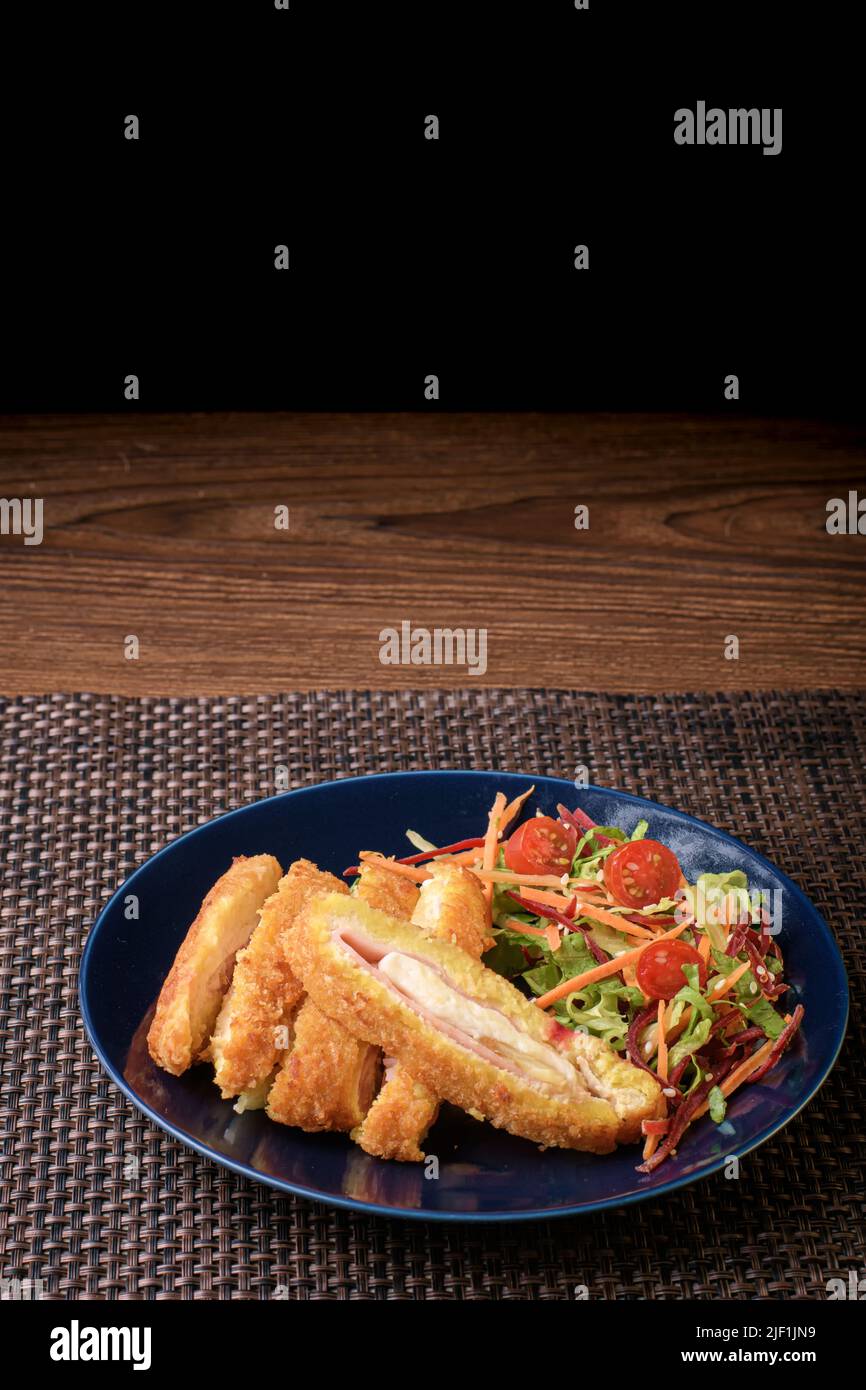 Köstlicher Salat aus grünem und rotem Gemüse, begleitet von panierten Hähnchen, gefüllt mit Schinken und Käse auf einem dunklen Teller Stockfoto
