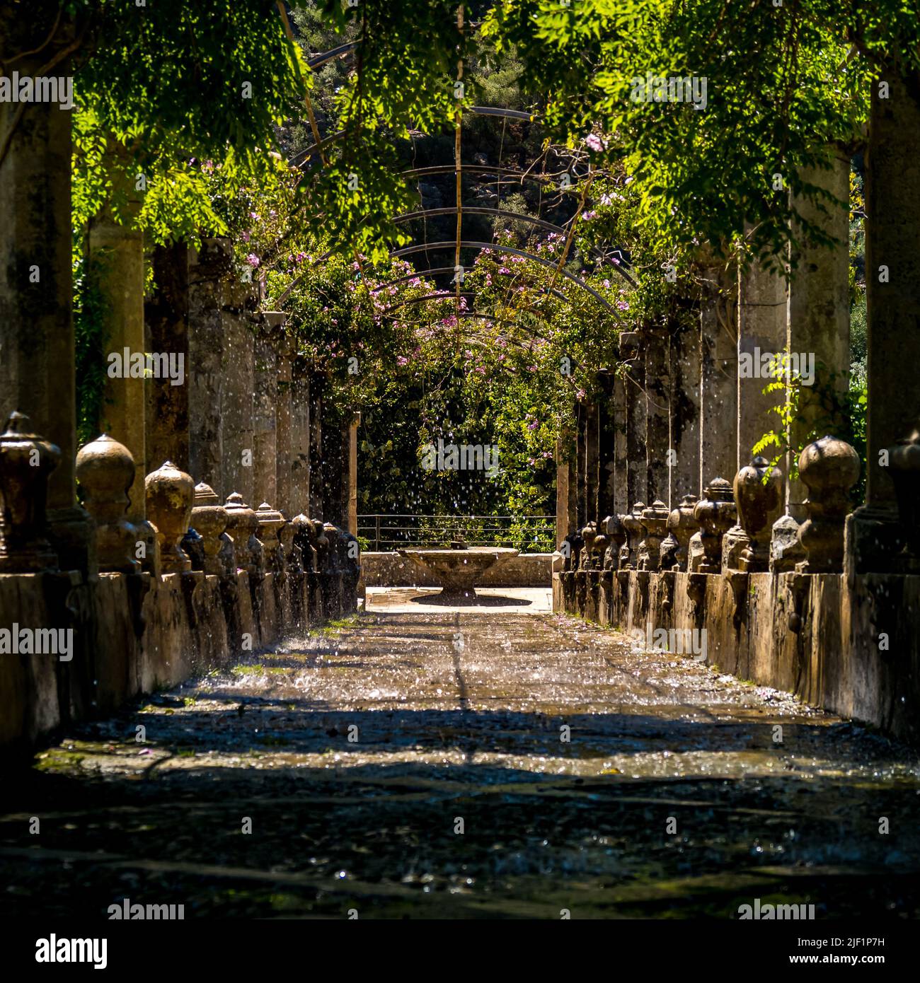 Ein nasser Pfad, der unter einer mit Steinsäulen gesäumten Pergola von Sprinklern überflutet wird, führt zu einem Brunnen mit berühmten Mallorca Alfabia Gärten. Stockfoto
