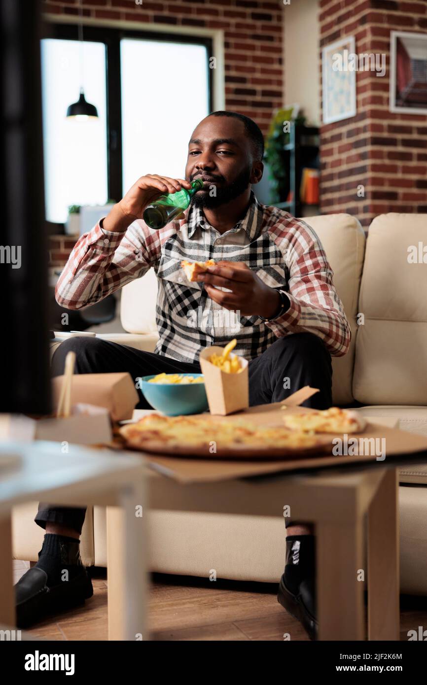 Glückliche Person trinkt eine Flasche Bier und isst Essen zum Mitnehmen von Fast-Food-Lieferung. Genießen Sie Essen zum Mitnehmen und schauen Sie sich Filme im Fernsehprogramm zu Hause an, essen Sie zu Abend und trinken Sie. Stockfoto