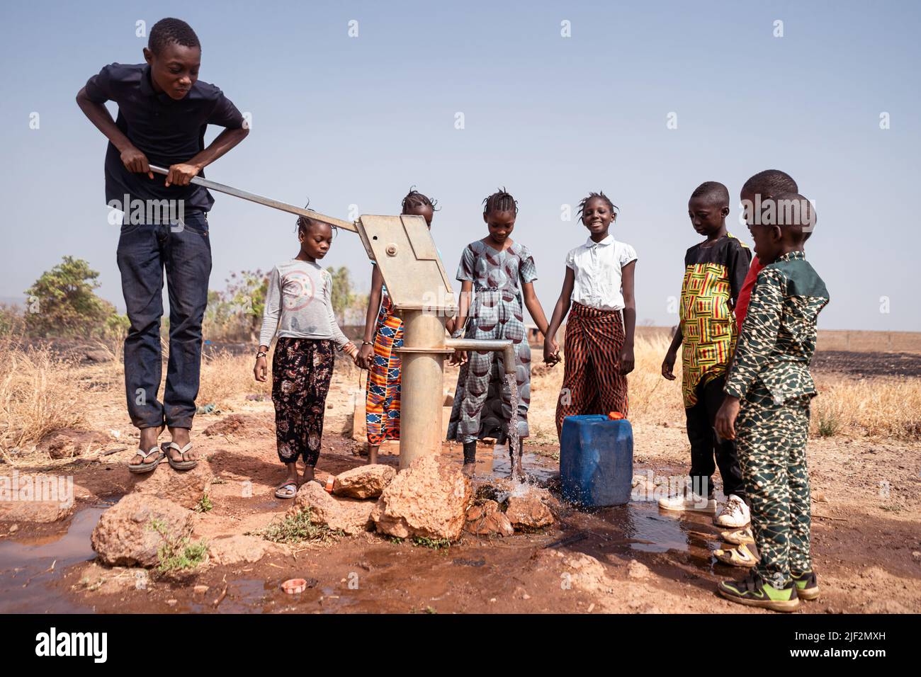 Mehrere junge afrikanische Dorfkinder gruppierten sich um eine manuelle Pumpe, die Kanister mit sauberem und frischem Trinkwasser füllt Stockfoto