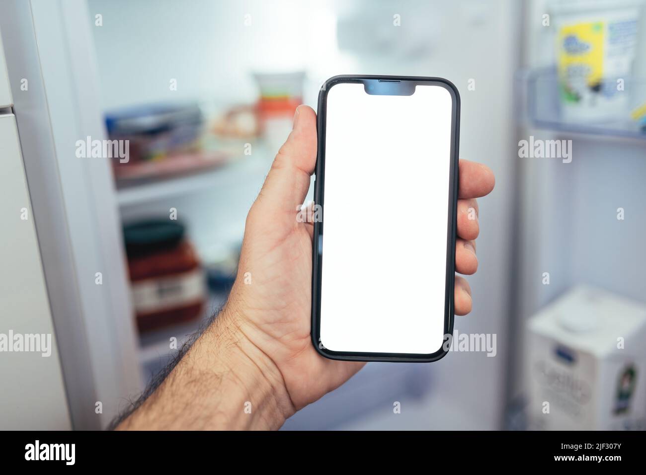 Smartphone-Modell für das Internet der Dinge, ein Mann hält ein Smartphone mit leerem Bildschirm vor dem Kühlschrank und überprüft die zusatzkosten für Lebensmittel Stockfoto