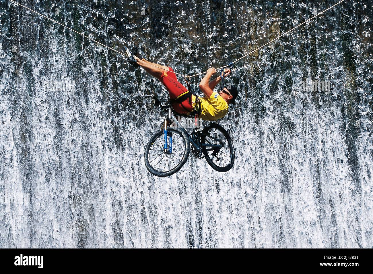 Mountainbiker mit seinem Fahrrad überqueren einer Schlucht hängen an einem Kletterseil, Wasserfall im Hintergrund Stockfoto