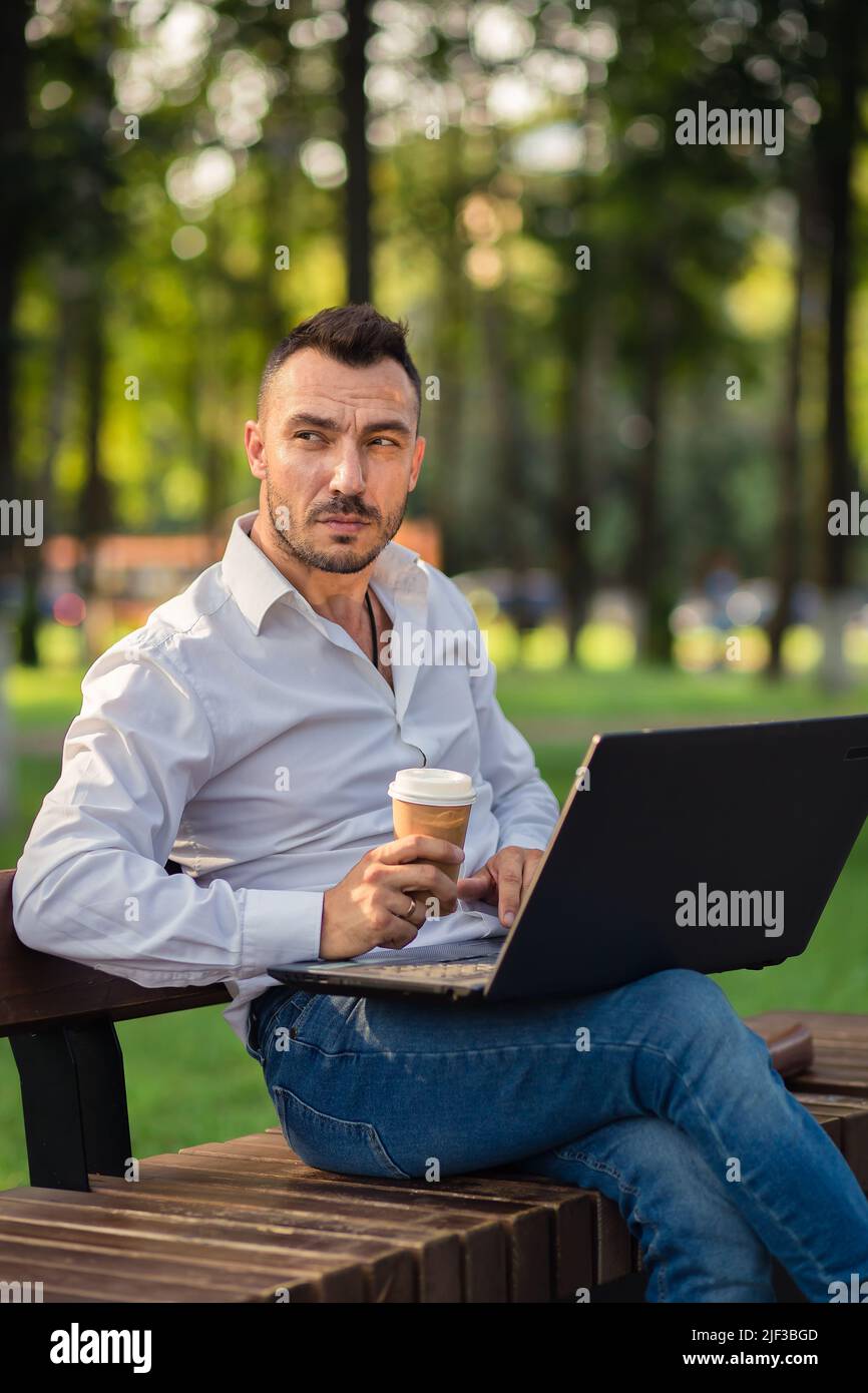 Serious Mann arbeitet im Park mit einem Laptop, trinkt Kaffee. Ein junger Mann auf einem Hintergrund von grünen Bäumen, ein heißer sonniger Sommertag. Warmes, weiches Licht, Nahaufnahme. Stockfoto