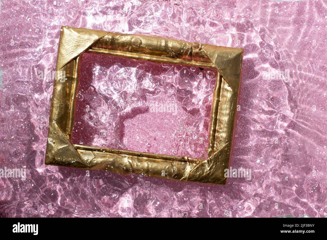 goldener Rahmen, der auf die Wasseroberfläche spritzt, mit sandigem rosa Glitzer auf dem Boden. Stockfoto