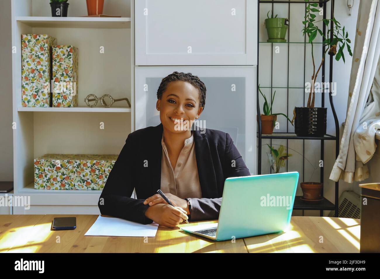 Lächelnd Junge Erwachsene Unternehmer freiberufliche schwarze Frau Kleinunternehmer mit Laptop und Smartphone arbeiten im Home Office Blick auf Kamera aufgenommen Stockfoto