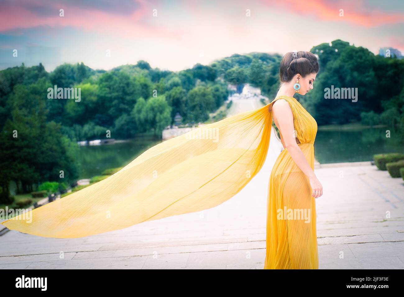Schönes weibliches Modell mit langem gelben Seidenkleid, das wie Flügel im Wind fliegt und im Profil auf der Treppe im Park steht. Panoramafenster Stockfoto