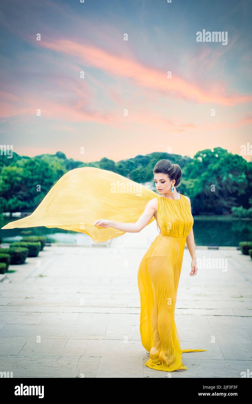 Schönes weibliches Modell mit langem gelben Seidenkleid, fliegend im Wind wie Flügel, stehend im Halbprofil auf der Treppe im Park. Panorami Stockfoto