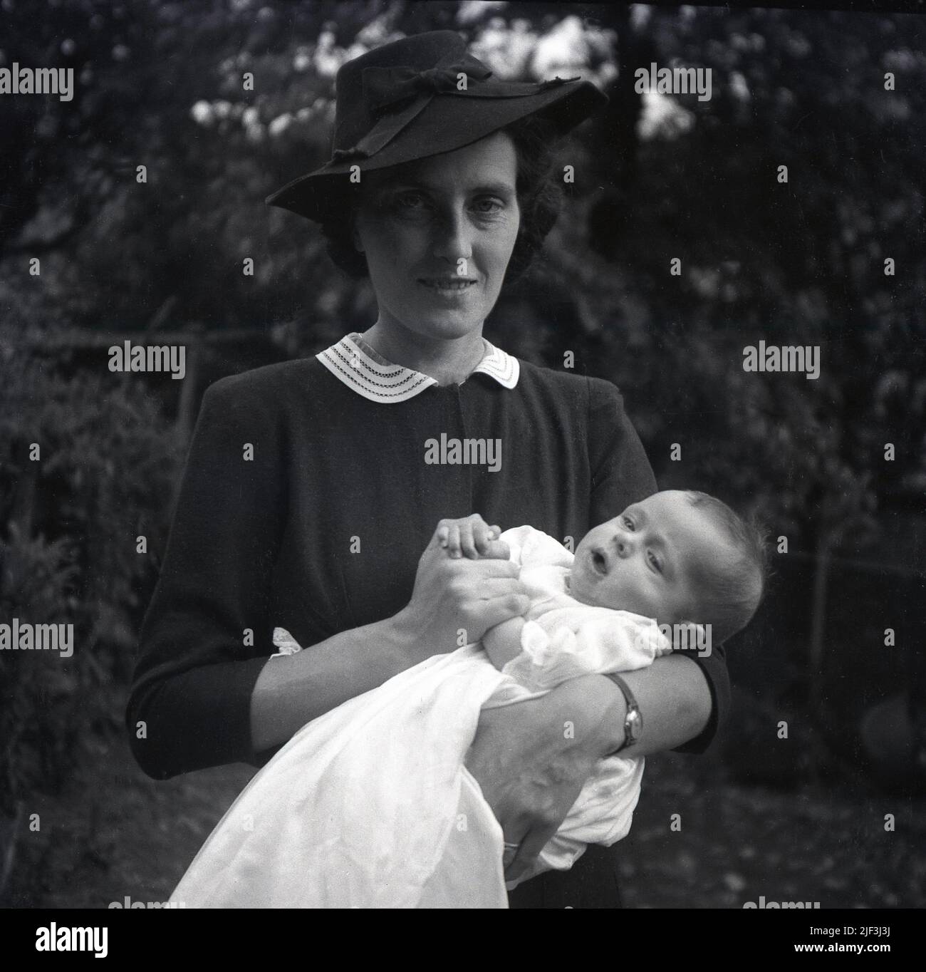 1950s, historisch, draußen in einem Gartenbereich stehend, eine elegant gekleidete Dame, die einen Hut trägt und ihr frisch getauftes Baby in der Hand hält, England, Großbritannien. Stockfoto