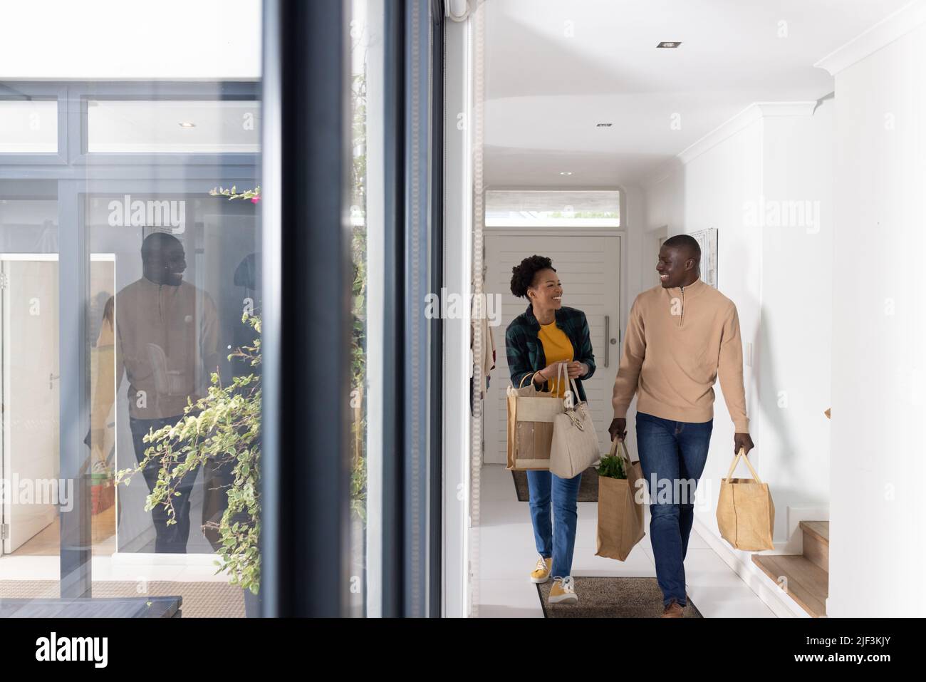 Glückliches afroamerikanisches Paar mit mittleren Erwachsenen und Einkaufstaschen, das zu Hause auf dem Flur läuft Stockfoto