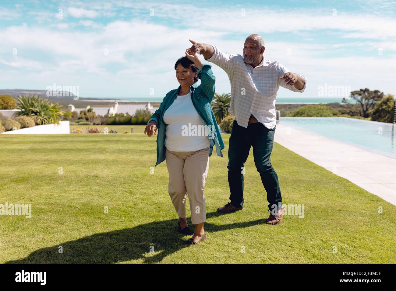 Glücklicher älterer Biracial Mann, der im Sommer mit der Frau auf grasbewachsenem Land gegen den Himmel im Hof tanzt Stockfoto