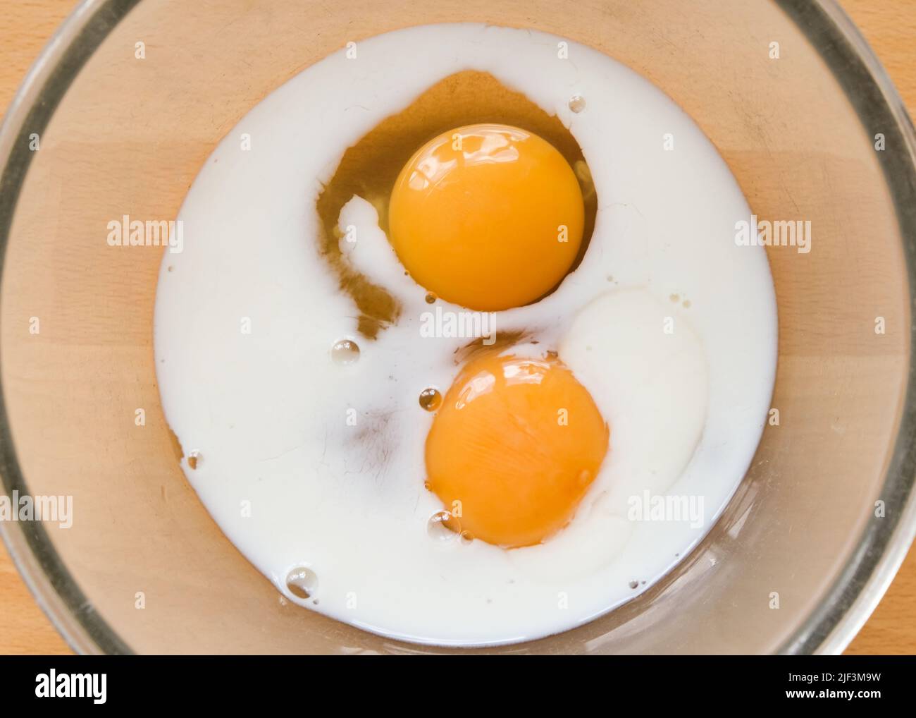 Milch, die zwei rohen Hühnereiern Eigelb in einer Glasschüssel zugesetzt wird, bereit, Rührei zu machen. Ei- und Milchmischung Stockfoto