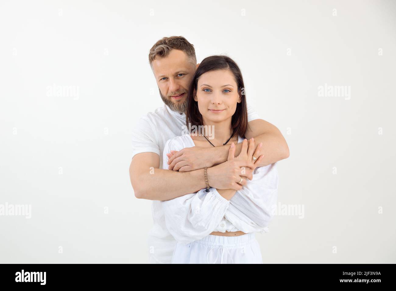 Bärtiger Mann hinten und Frau, glückliche Familie in hellem Outfit stehen zusammen, schauen auf die Kamera, umarmt auf weißem Hintergrund. Stockfoto