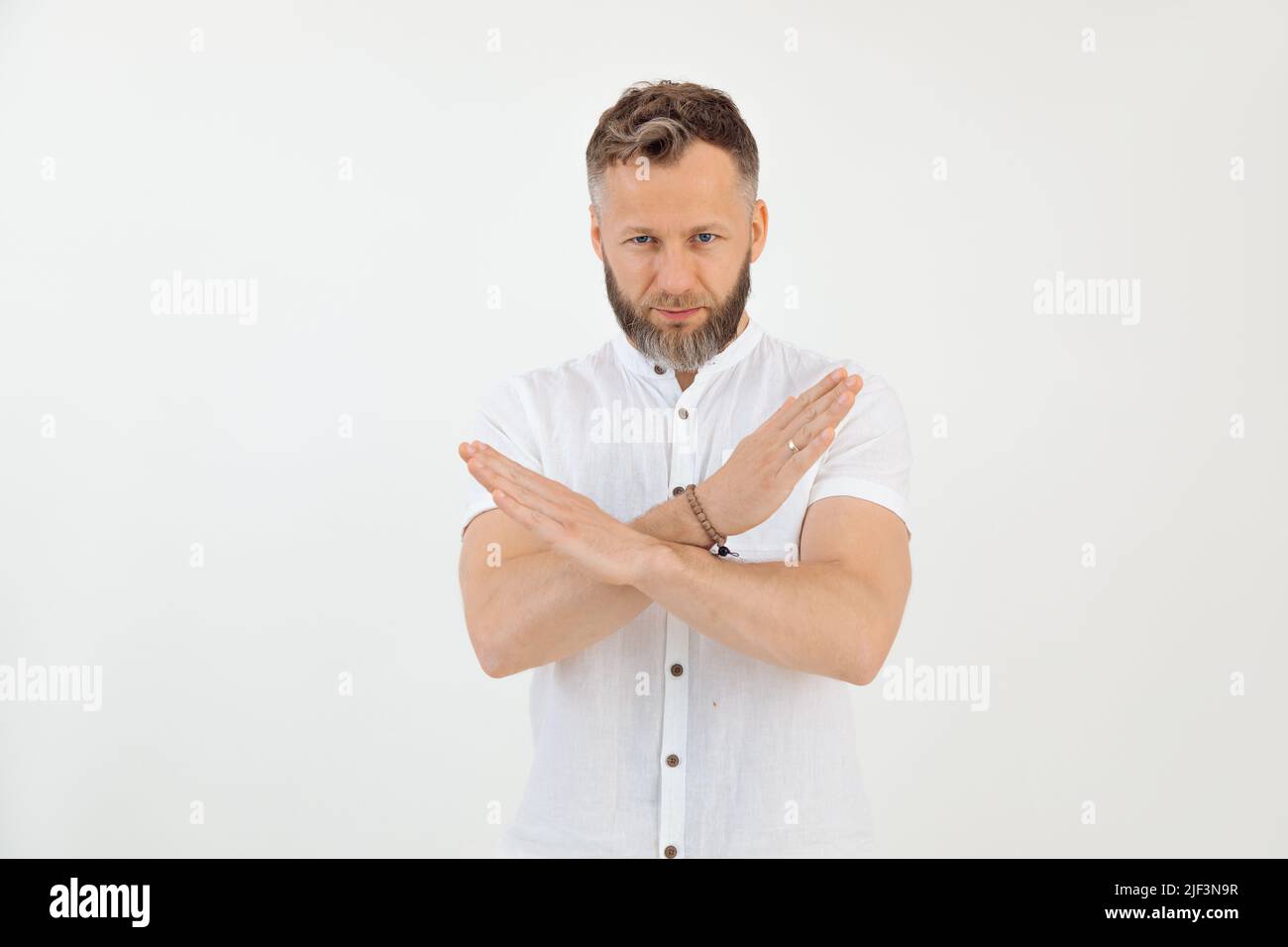 Bärtig ernster tätowierter Mann, der in weißem Outfit mit gekreuzten Händen negativ auf die Kamera schaut, leugnet, lehnt ab und schränkt ein Stockfoto