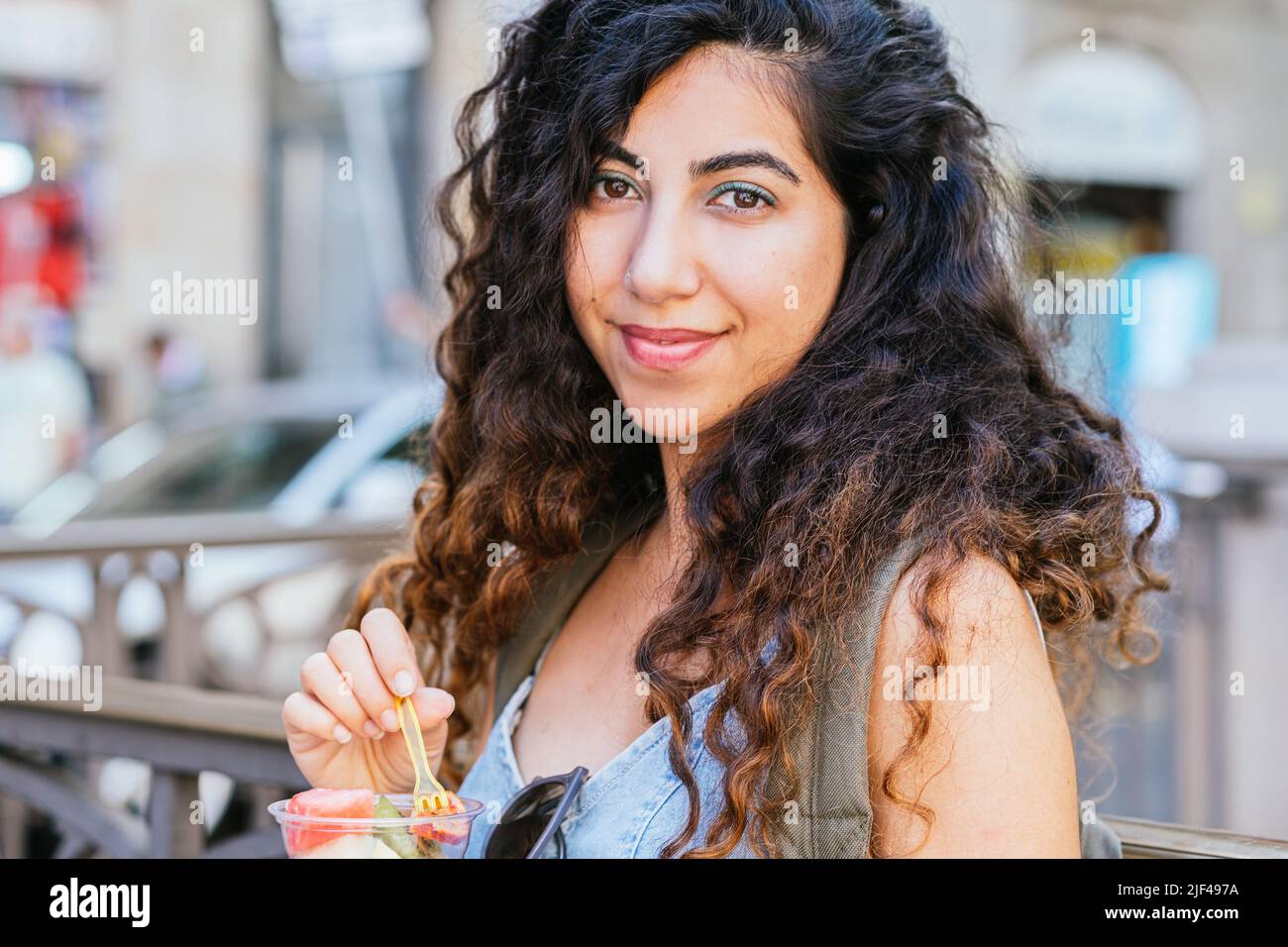 Junge dunkelhaarige Frau, die die Kamera anschaut, während sie einen Plastikbecher mit Fruchtstücken in der Hand hält Stockfoto