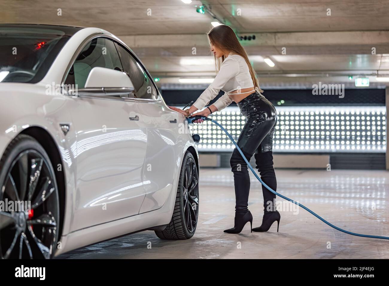 Attraktive junge Frau lädt ein Luxus-Elektroauto an einer Ladestation für Elektrofahrzeuge in einer öffentlichen Garage Stockfoto