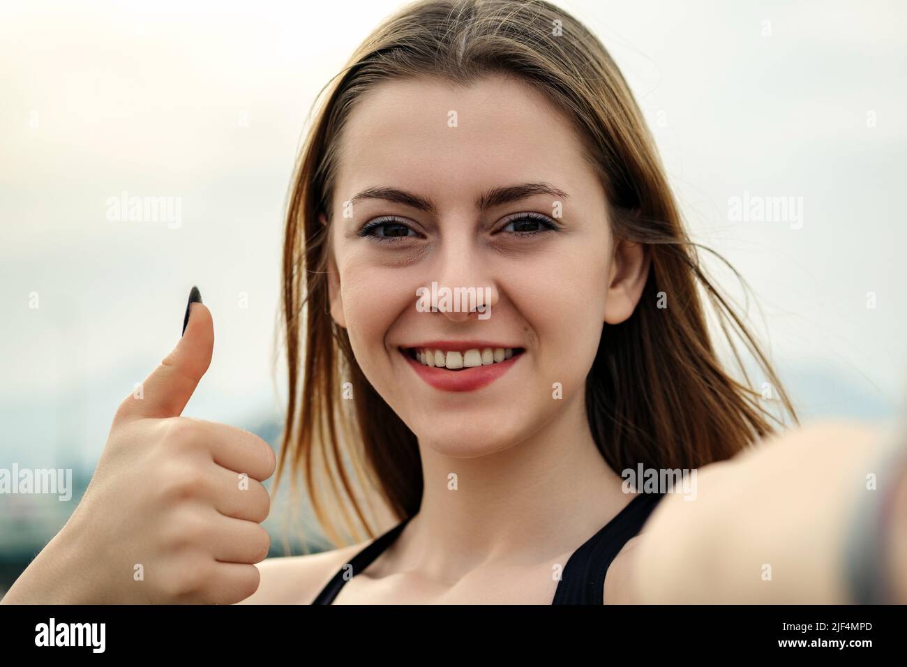 Nette sportliche Frau in einem schwarzen Sport-BH, die im Stadtpark steht und im Freien den Daumen hochzeigt und Selfie-Fotos macht. Blick auf die Kamera. Stockfoto