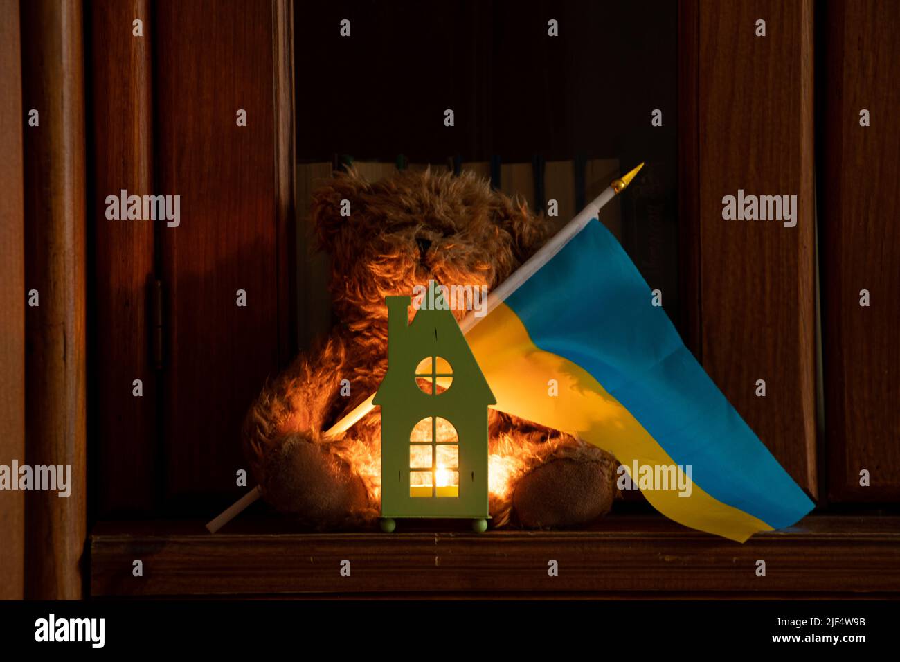 Ein kleines Kinderhaus und eine Kerze daneben liegt ein Teddybär mit der Flagge der Ukraine auf den Regalen zu Hause, der Krieg in der Ukraine, Heimat Ukraine Stockfoto