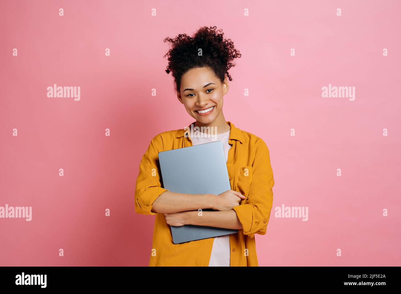 Schöne positive afroamerikanische junge Frau mit lockigem Haar, in lässiger, stilvoller Kleidung, mit Laptop, über einem isolierten rosa Hintergrund stehend, mit Blick auf die Kamera, freundlich lächelnd Stockfoto