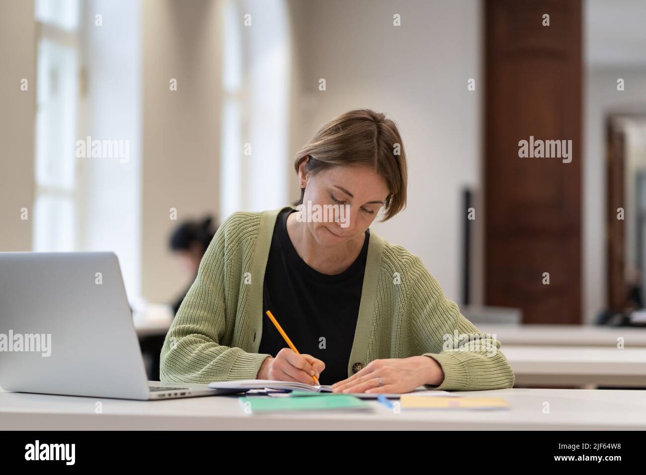 Studentin mittleren Alters, die sich auf die Prüfung in der öffentlichen Bibliothek vorbereitet, während sie mit einem Laptop am Tisch sitzt Stockfoto