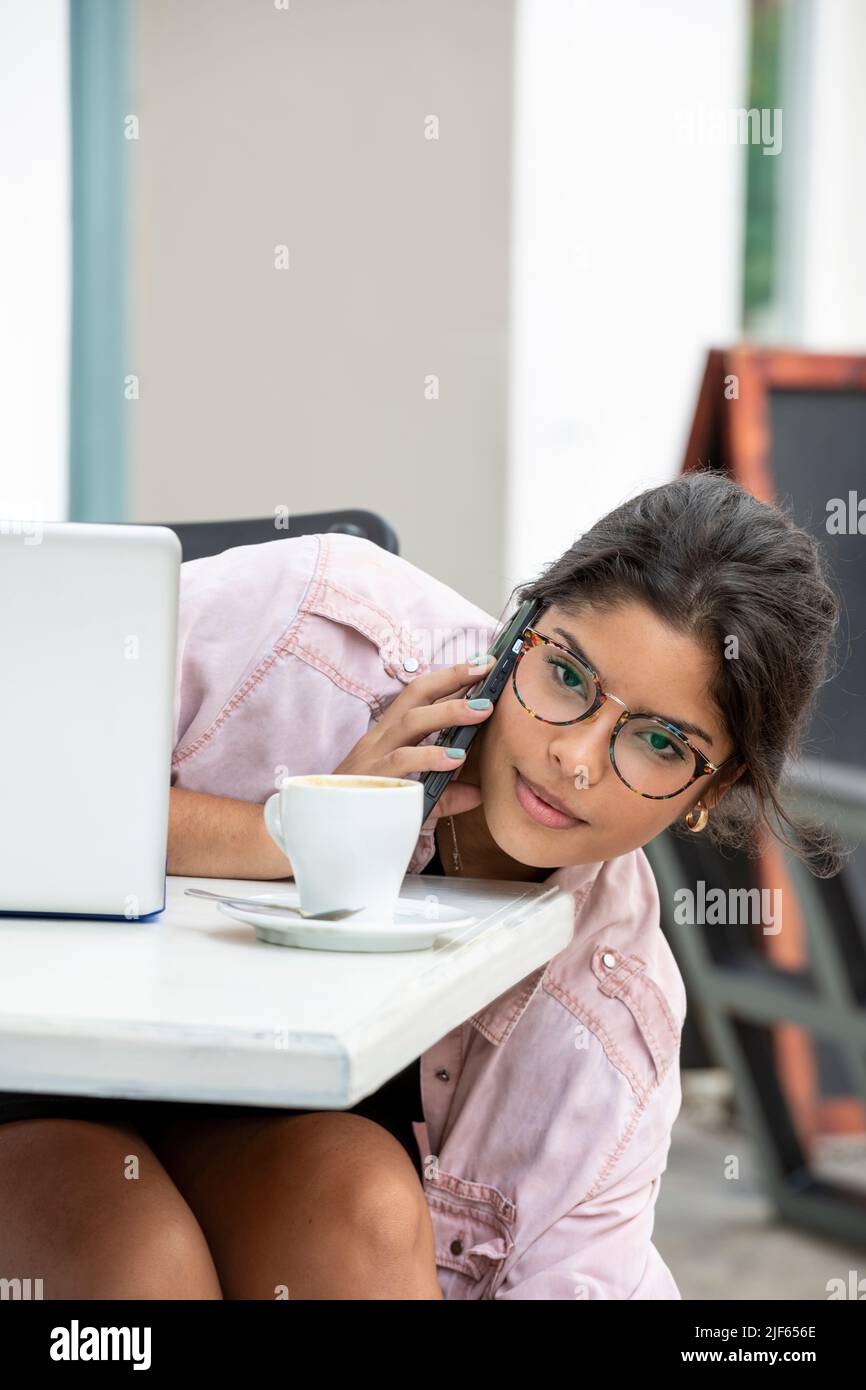 Lateinische junge Frau mit Laptop und Telefon, während sie im Straßencafé sitzt - Stockfoto Stockfoto