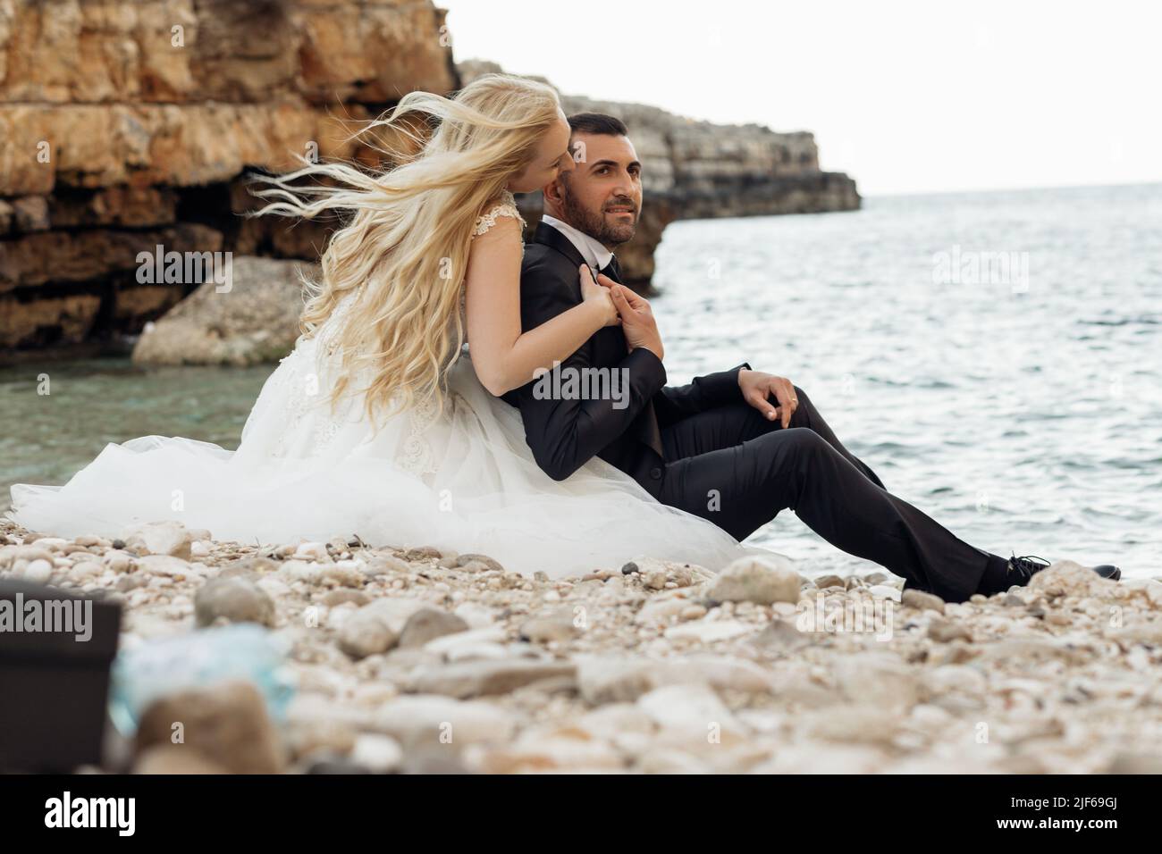 Seitenansicht eines wunderschönen Paares, das auf Steinkieseln am Ufer in der Nähe des Wassers sitzt. Junge Frau Braut umarmt küssen Bräutigam. Stockfoto