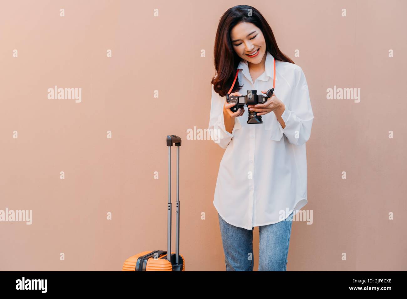Junge alleinreisende asiatische Frau, die die Fotos aus ihrem Urlaub vor der Kamera mit einer sandfarbenen Wand im Hintergrund und einem orangefarbenen Koffer neben ihr in legerer Kleidung mit Platz für Kopien überprüft Stockfoto