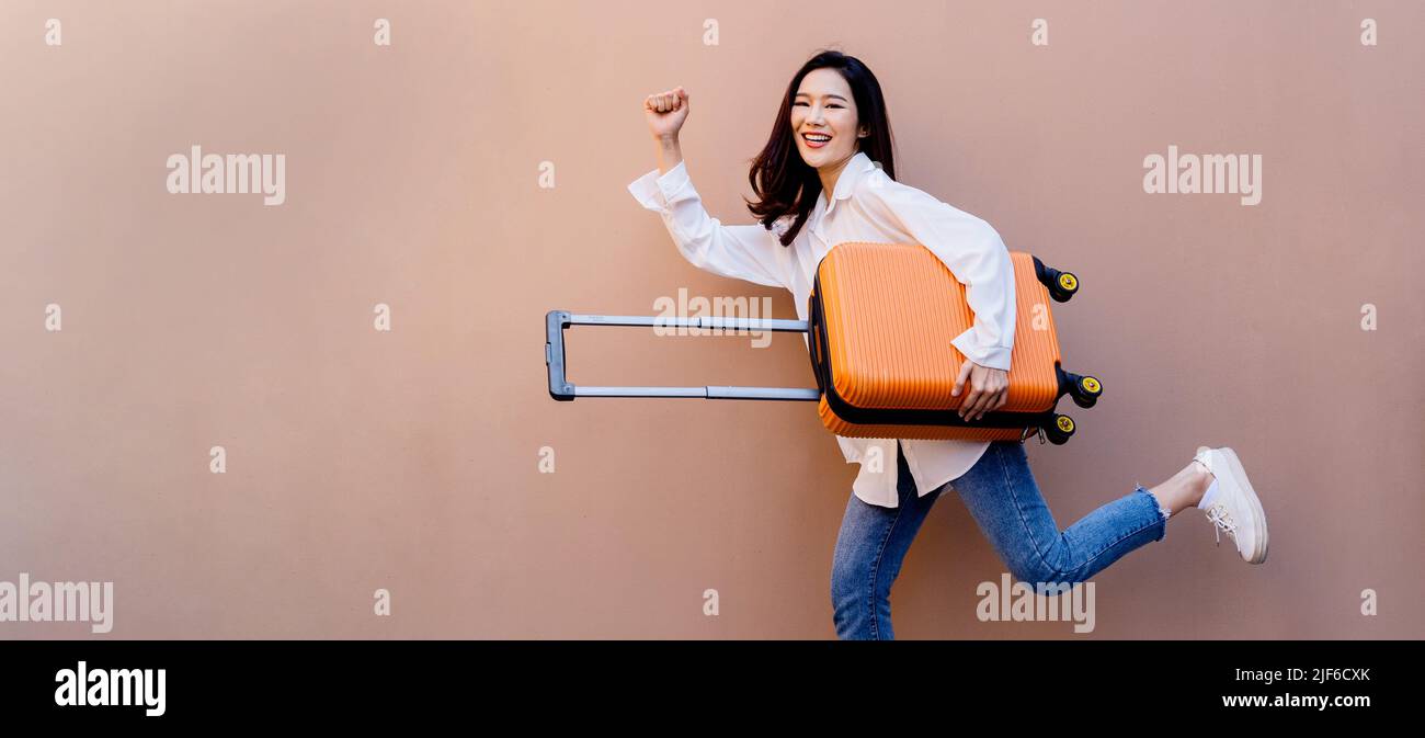 Junge asiatische Mädchen glücklich, während Schleppen ihre leuchtend orange Koffer mit der rechten Hand und der linken Hand Faust stoßen in der Luft auf Betonboden und Wüste Sand farbige Wand in ihrem Hintergrund. Stockfoto
