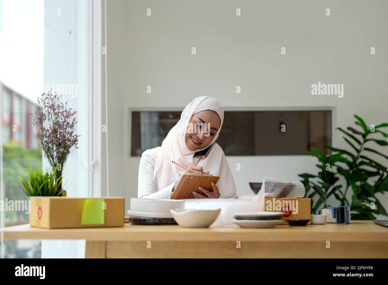 Junge asiatische muslimische Frau Geschäftseigentümer mit Paketboxen Online-Verkauf Job, Anruf mit Smartphone, erhalten Sie eine Bestellung von Kunden, Notizen zu machen, und machen Stockfoto