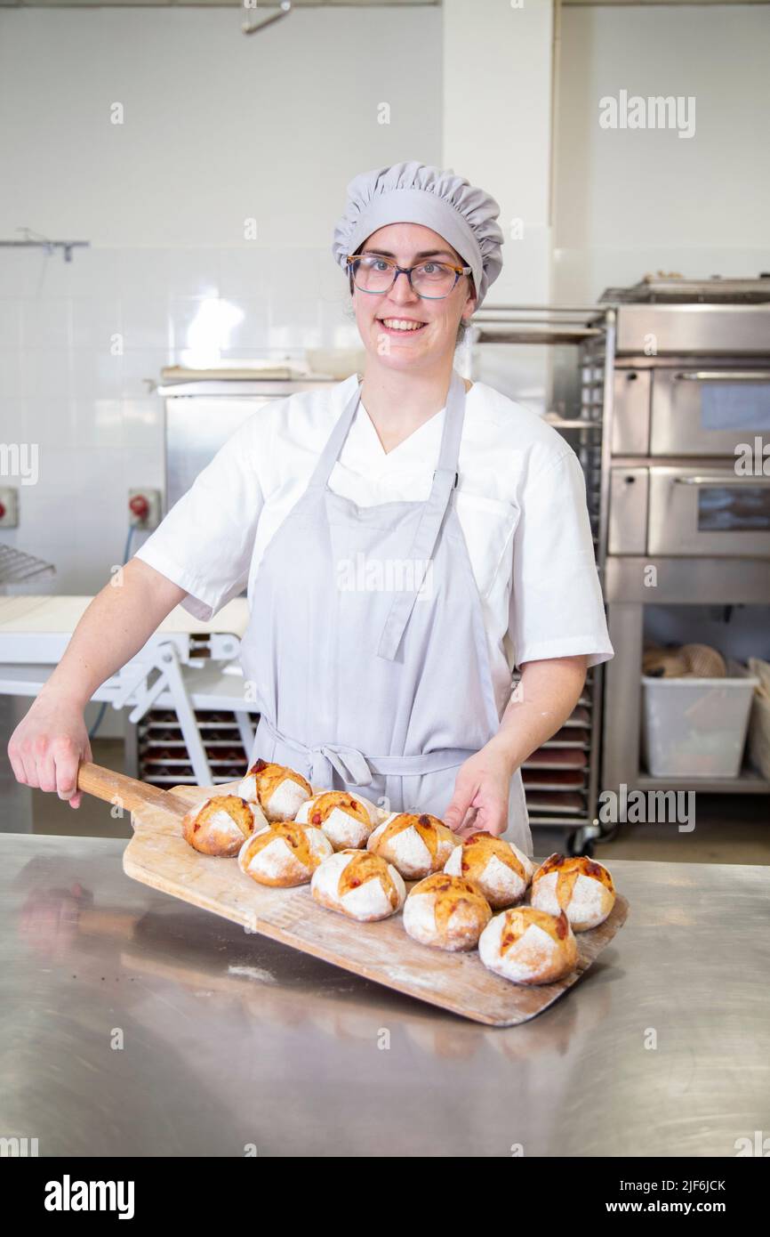 Fröhliche Bäckerin in Uniform mit Fülle von kleinen gebackenen Brot Blick auf Kamera am Tisch in der Bäckerei mit Mitarbeitern Stockfoto