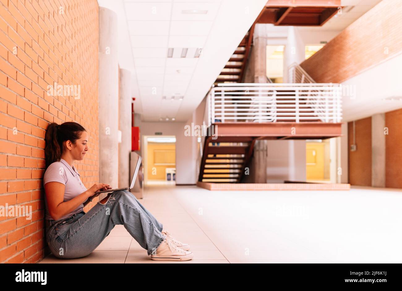 Ganzkörper-Seitenansicht einer entzückten Studentin in Freizeitkleidung, die auf Netbook im Internet surft, während sie auf dem Boden in der Nähe einer Ziegelwand sitzt Stockfoto