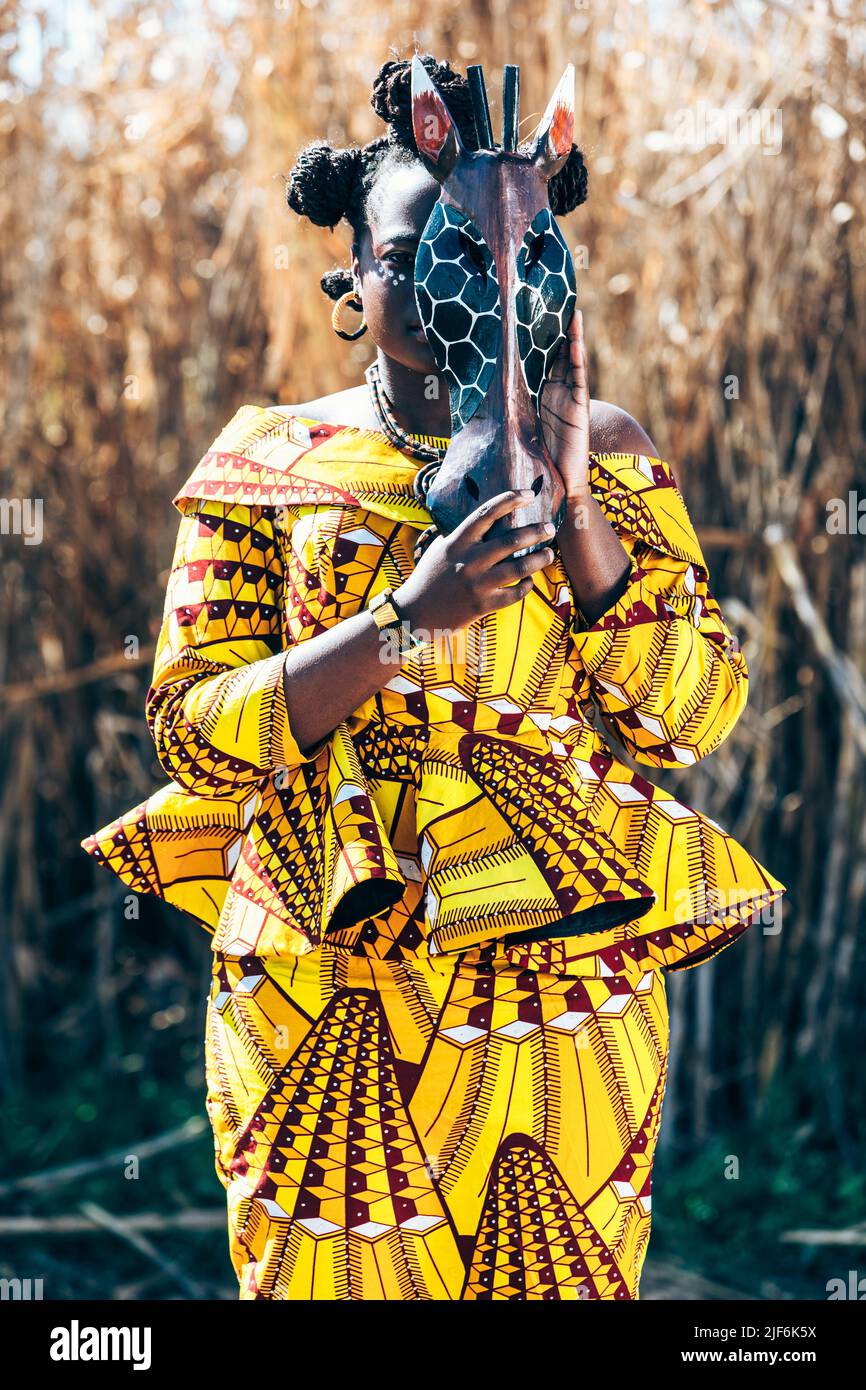 Afrikanische Frau in gelbem Kleid mit authentischem Aufdruck, das Gesicht mit Pferdemaske bedeckt, während sie in der Nähe von trockenem Gras auf dem Feld steht Stockfoto